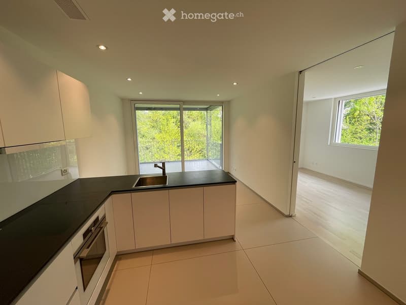 Modern kitchen with white cabinets, black countertops, and a sink, leading to a balcony with a view of greenery.