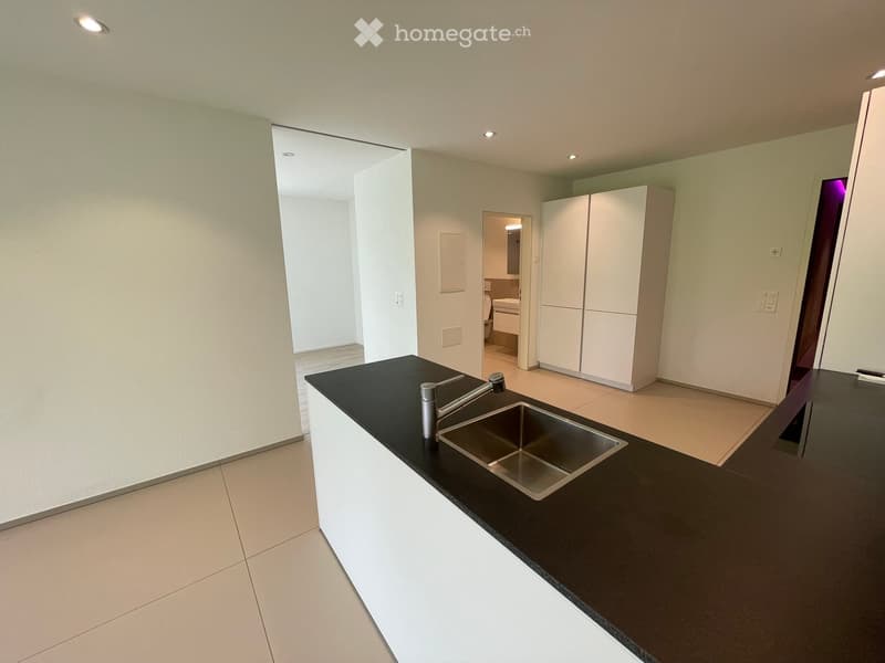 Modern kitchen with black countertop, stainless steel sink, and white cabinets. Open layout with visible bathroom area in the background.