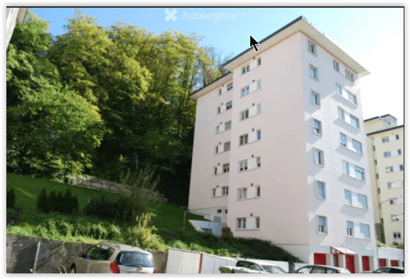 Multi-story residential building with light pink facade, multiple windows, and raised ground floor. Surrounded by greenery and parked cars.