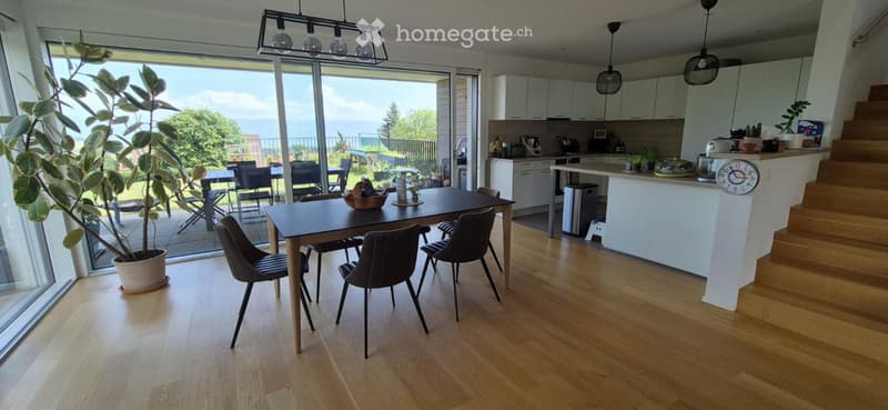 Bright, open-plan living and dining area with light wood flooring, large windows, a dining table with chairs, a kitchen island, and a potted plant near the window.