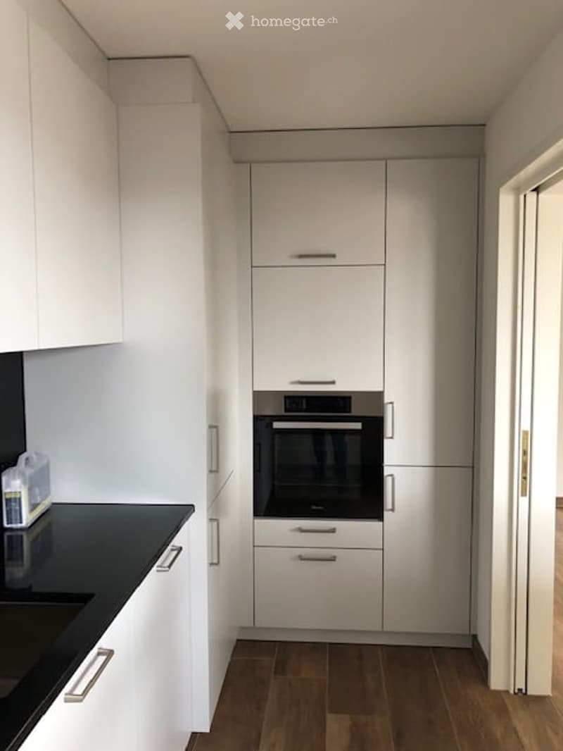 Modern kitchen with white cabinets, black countertop, and built-in oven. Wooden flooring and sleek design.