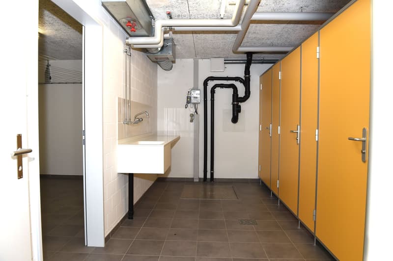 A bathroom with a white sink, shower cubicles, tiled floor, and metal pipes attached to the wall.