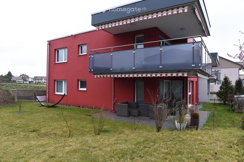 Two-story red house with a modern design, featuring a large balcony with glass railings and striped awnings, surrounded by a well-maintained lawn and patio seating area.