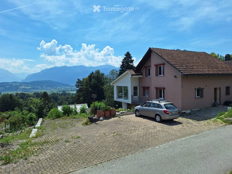 Two-story house with pink siding, tiled roof, and large windows, situated in a scenic mountainous area with a driveway and parked car.