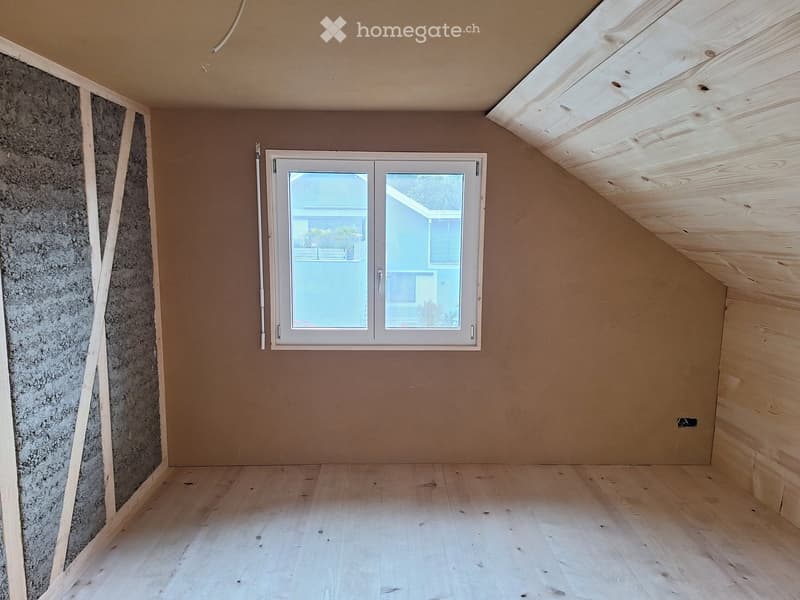 Empty attic room with wooden flooring, exposed wooden ceiling beams, and a single window with white frame. Walls are unfinished with one side showing insulation material.