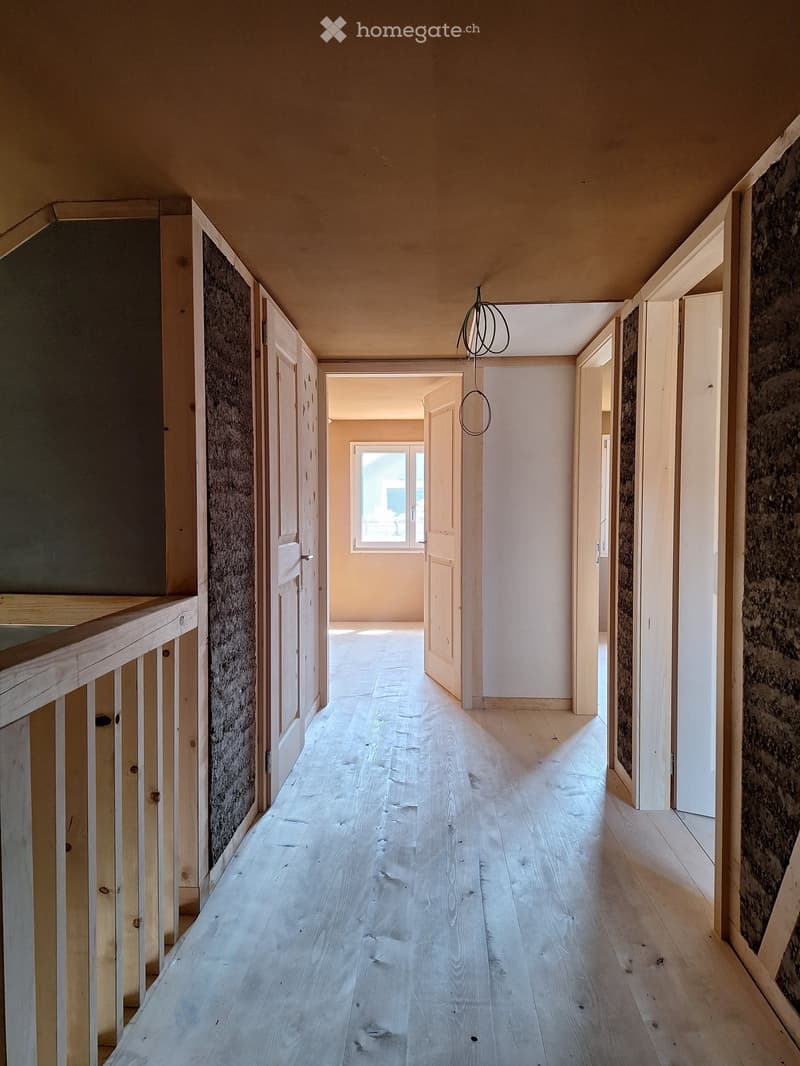 Empty hallway with wooden flooring, white walls, and several open doors leading to other rooms. Natural light streams in from a window at the end of the corridor.