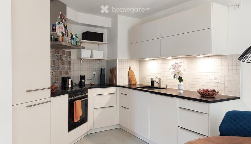 Modern kitchen with white cabinets, black countertops, stainless steel sink, and various appliances including an oven and coffee maker. Shelves hold decorative items and kitchen essentials.