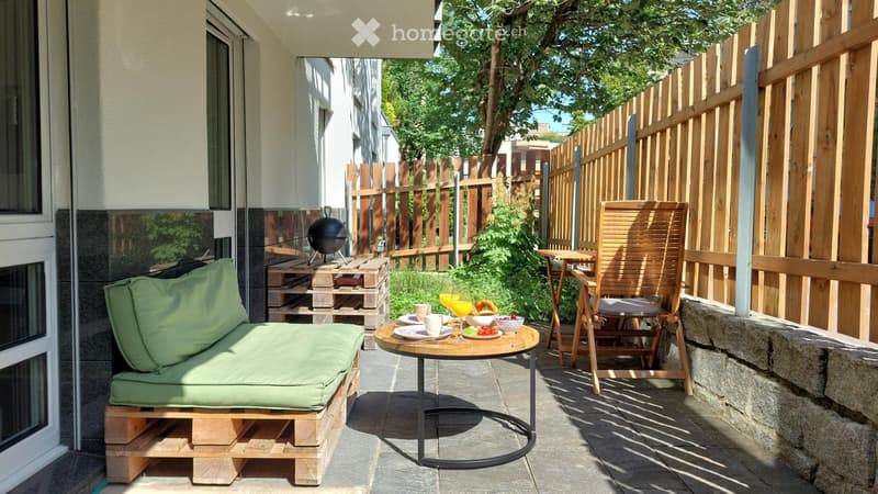 Balcony with a green sofa, small round table, wooden chair, and various items including cups, a glass of orange juice, and bowls of fruit.