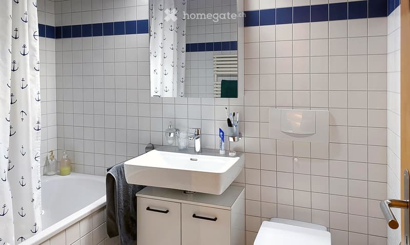 Modern bathroom with white tiles, a white sink, a bathtub, a mirror, and a toilet. The walls feature a blue accent border, and a towel hangs on the side of the sink.