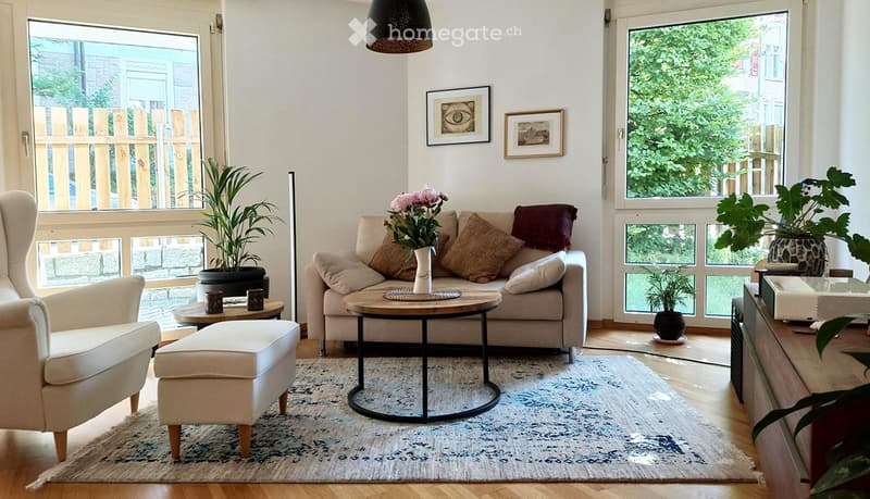 Modern living room with a grey sofa, coffee table, ottoman, armchair, and potted plants. Large windows offer a view of greenery outside.