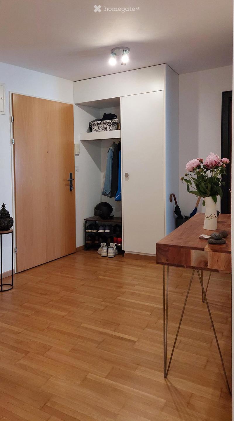A modern hallway with wooden flooring, a built-in closet with shelves and hanging space, a small table with a vase of flowers, and an umbrella stand.