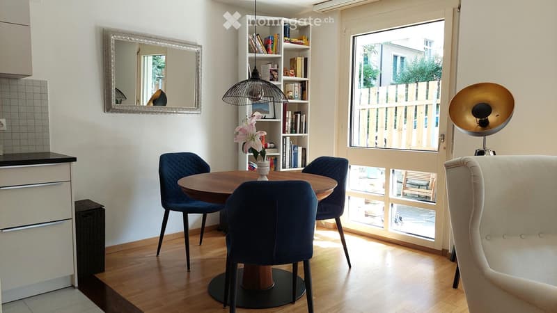 A modern, well-lit room with a round wooden dining table, four blue chairs, a white sofa, a bookshelf filled with books and decorative items, a large mirror, and a hanging pendant light.