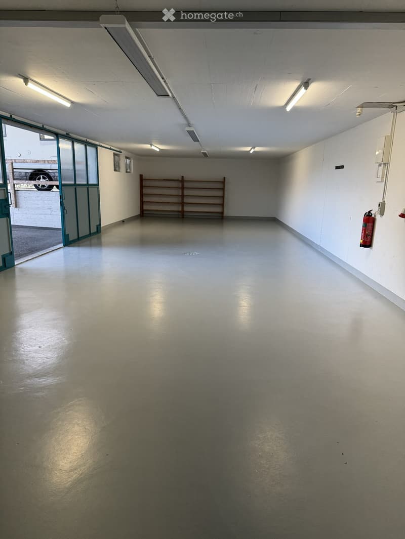 Empty garage with white walls, ceiling lights, wooden shelf, fire extinguisher, and an open door leading to another room.