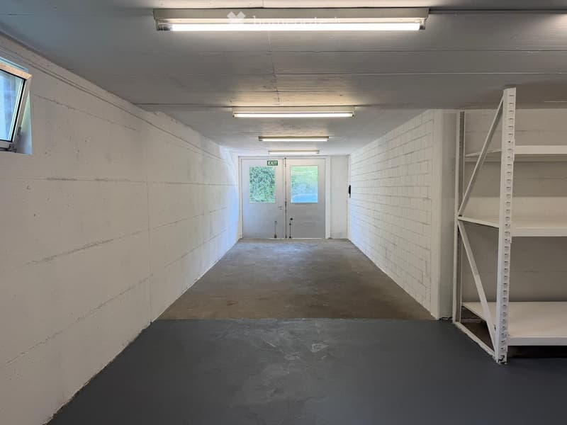Empty storage area with white brick walls, concrete floor, fluorescent lighting, and a metal shelving unit on the right side.