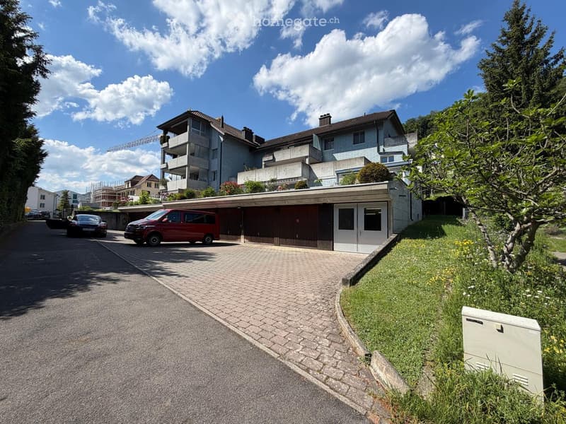 A modern multi-story house with a garage, surrounded by greenery and a paved driveway.