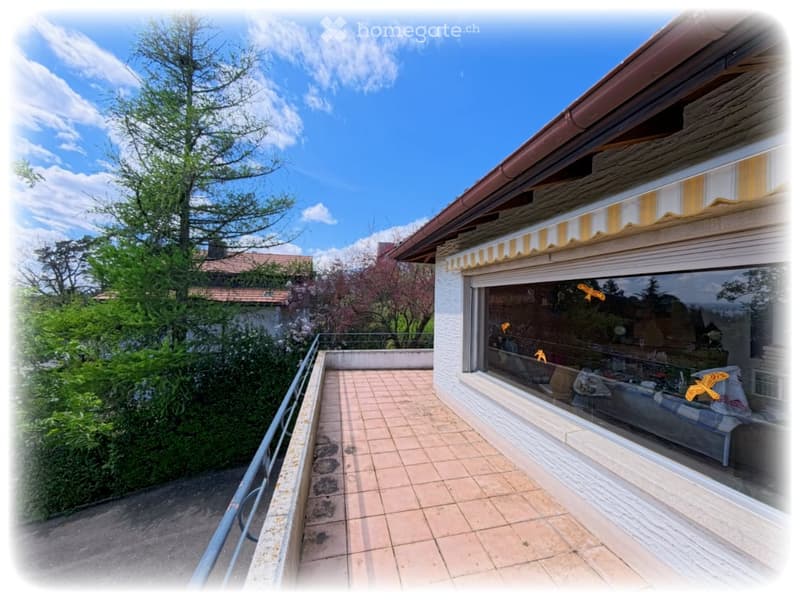 Balcony with tiled floor, metal railing, and striped awning above a large window. The view includes greenery and a clear blue sky.