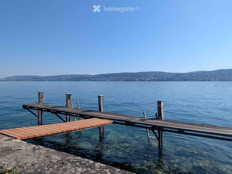 Wooden dock extending over calm water with a distant view of a town and hills under a clear blue sky.