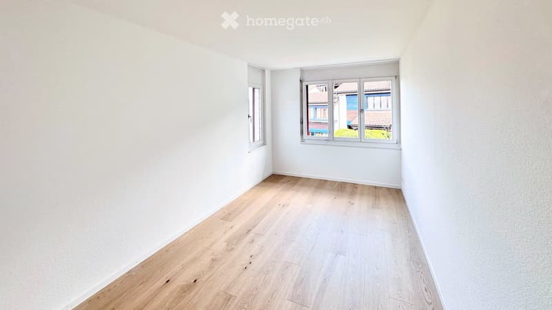 Empty room with white walls and light wooden flooring, featuring two windows with white frames and blinds.