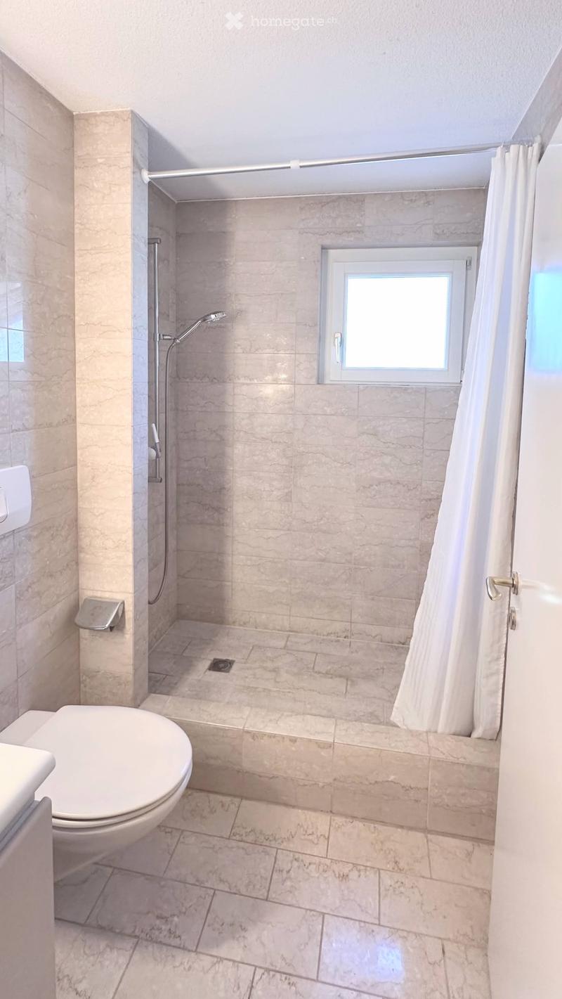 Modern bathroom with marble tiles, a toilet, a shower with a curtain, and a small window above the shower area.