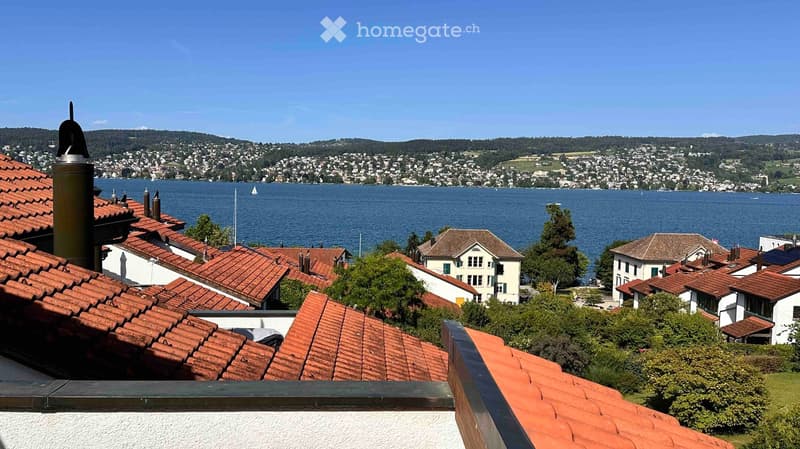 View from a balcony showing a lake, distant cityscape, and surrounding hills under a clear blue sky.