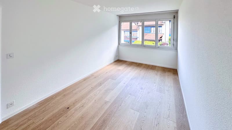 Empty room with light wooden flooring and white walls, featuring a large window with a view of neighboring buildings.