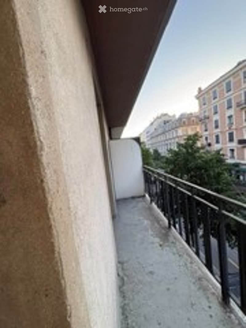 Balcony with metal railing, concrete floor, view of buildings and trees in the distance.
