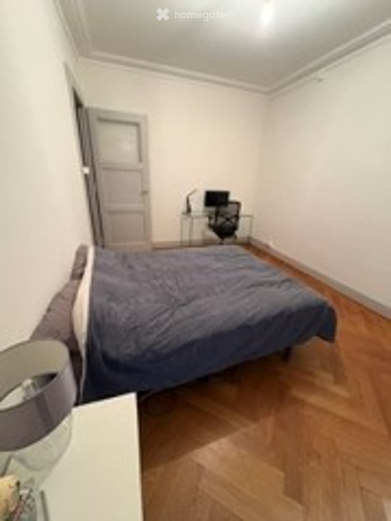 Bedroom with wooden flooring, a neatly made bed, a nightstand with a lamp, a small desk with a chair, and a closed door.