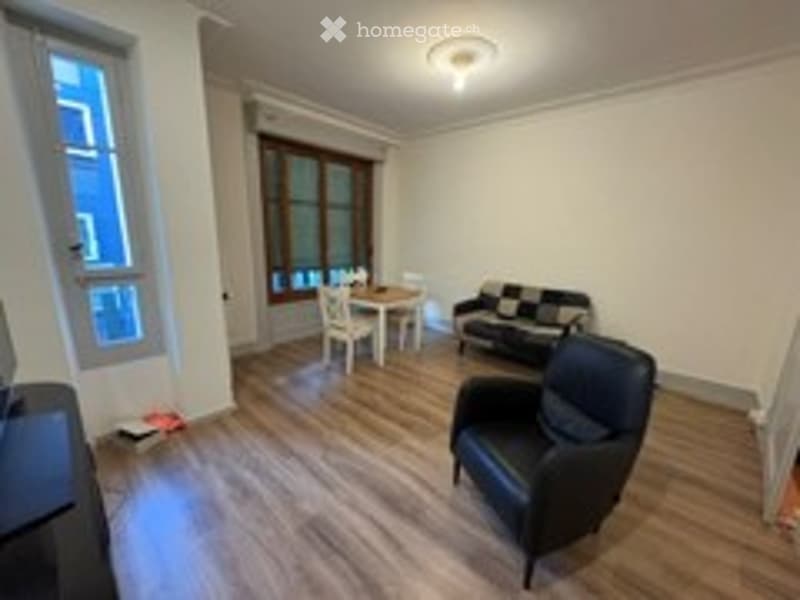 Bright, unfurnished living room with wooden flooring, large windows, a black sofa, a dining table with chairs, and an armchair.