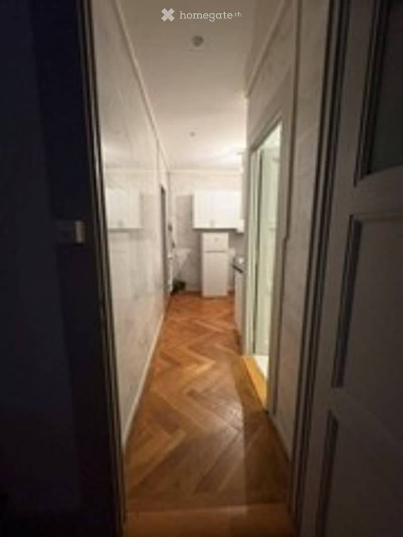 A narrow hallway with wooden flooring and white walls, leading to a room with a visible kitchen area.