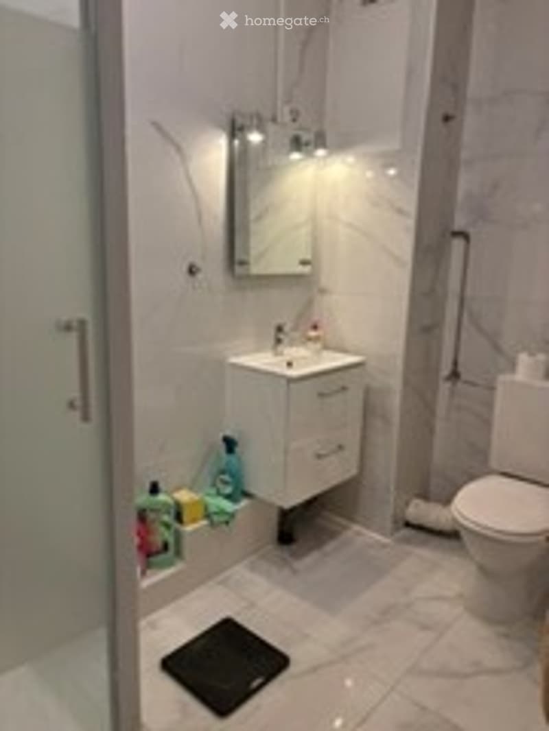 Modern bathroom with white tiles, a sink with a mirror, a toilet, a shower with a glass door, and cleaning supplies on the floor.