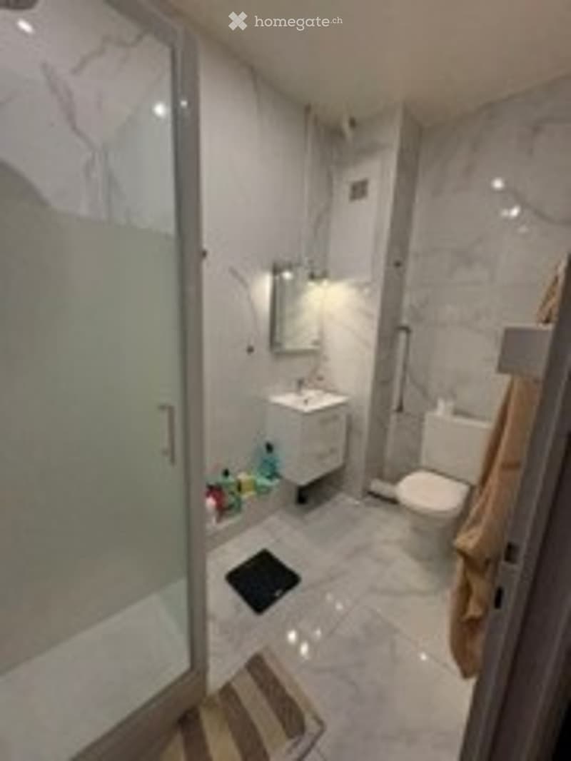 Modern bathroom with a white toilet, sink, and glass-enclosed shower. The walls are tiled in a marble-like pattern, and a towel hangs on a rack.