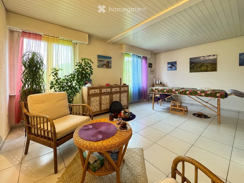 A well-lit room with a wicker sofa, a small wicker table with a purple plate, a wooden cabinet, a massage table, and several potted plants. The room features colorful curtains and artwork on the walls.
