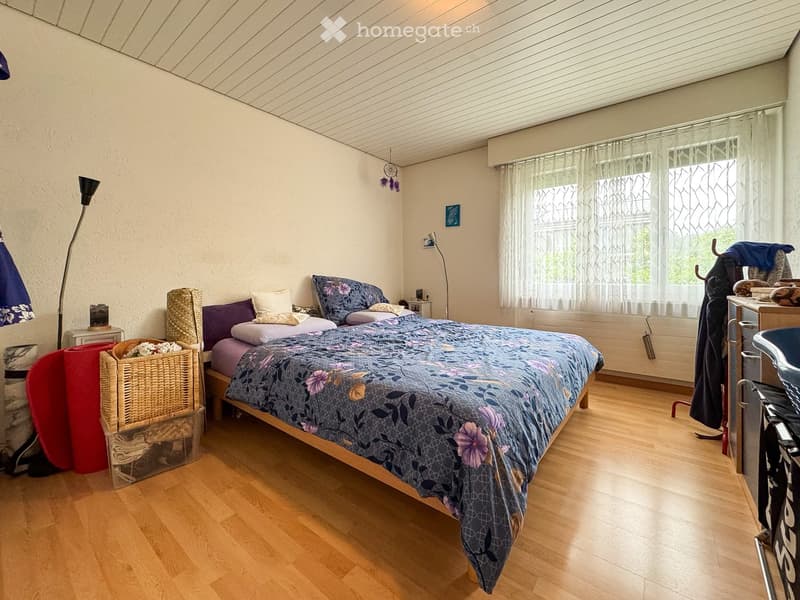 Cozy bedroom with wooden flooring, a large bed with floral bedding, a nightstand, a wicker basket, and a floor lamp. The room features large windows with sheer curtains and a slanted ceiling.