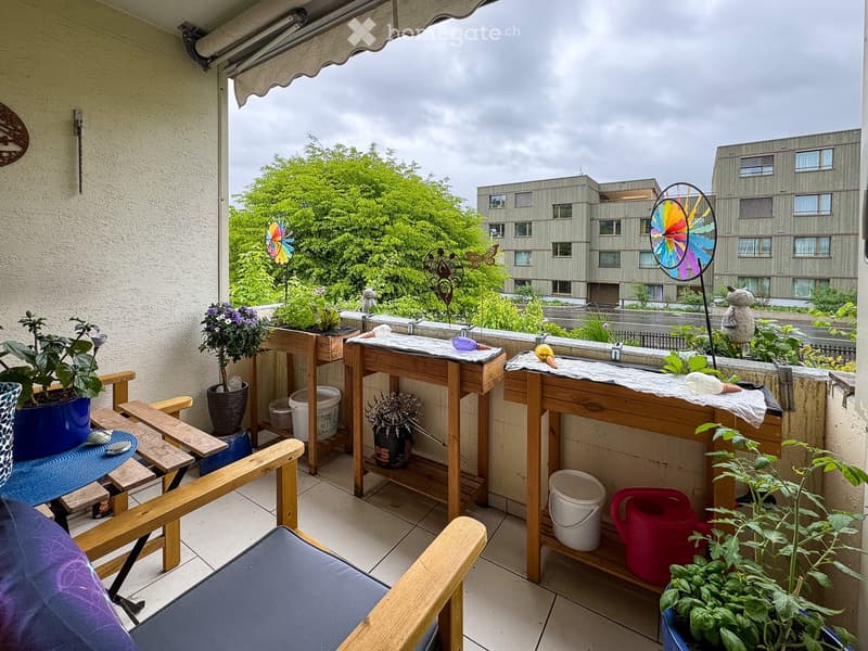 Balcony with wooden tables, potted plants, a chair, and garden tools, overlooking a building and greenery.