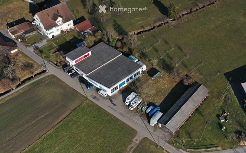 Aerial view of a rural building complex with a large industrial-style building, several parked vehicles, and surrounding fields.