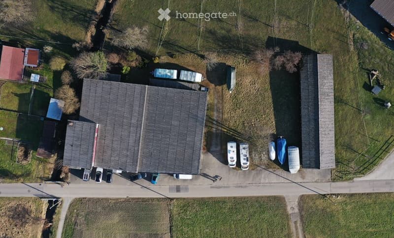 Aerial view of a large building with a flat roof, surrounded by smaller structures and parked vehicles, including cars and boats.