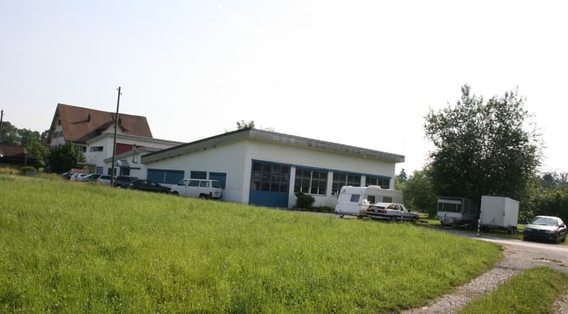 A single-story building with a flat roof and blue trim around the windows, surrounded by a grassy area with several parked cars and trailers.