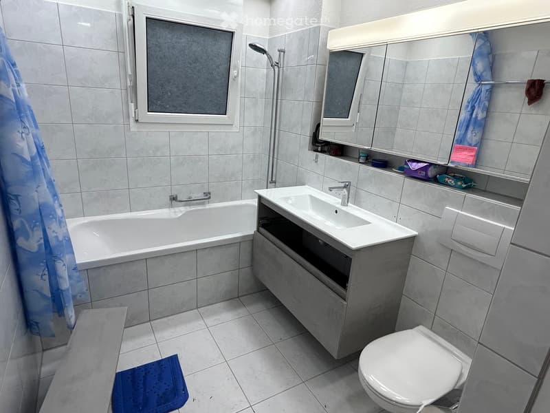 Modern bathroom with a bathtub, sink, toilet, mirror, and showerhead. The room features tiled walls and floor, a window, and a small shelf with toiletries.