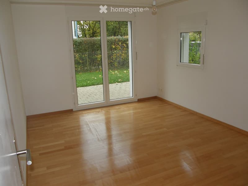 Empty room with wooden flooring, large sliding glass doors leading to a garden, and a single window on the opposite wall.