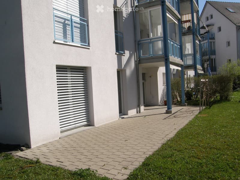 Modern multi-story residential building with white walls, blue-framed windows, and balconies. The entrance features a paved walkway leading to a door, with green grass and small plants on either side.