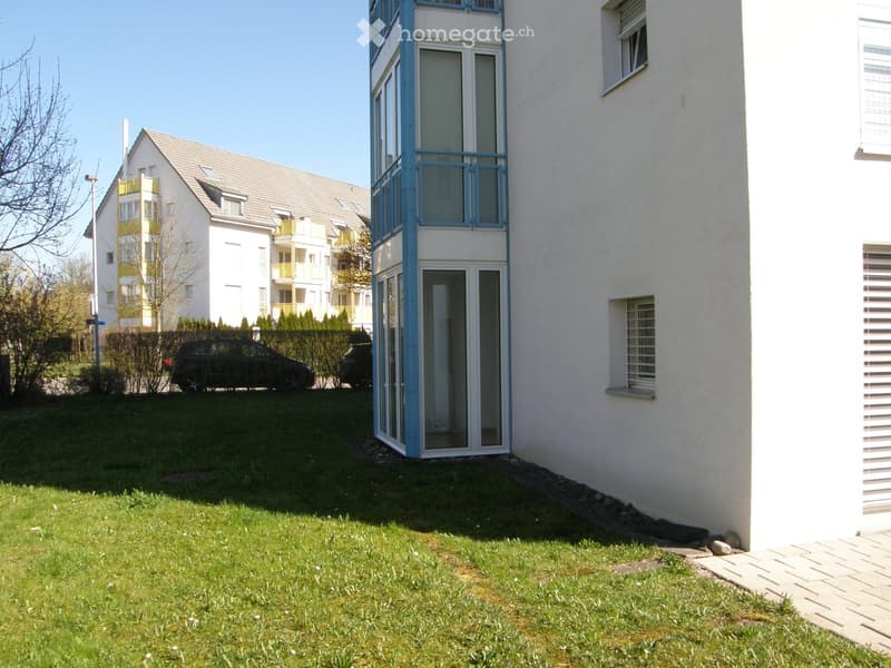 Modern multi-story residential building with white exterior, blue accents, and a raised ground floor entrance. The building features large windows and balconies, surrounded by a well-maintained lawn and a parking area with cars.
