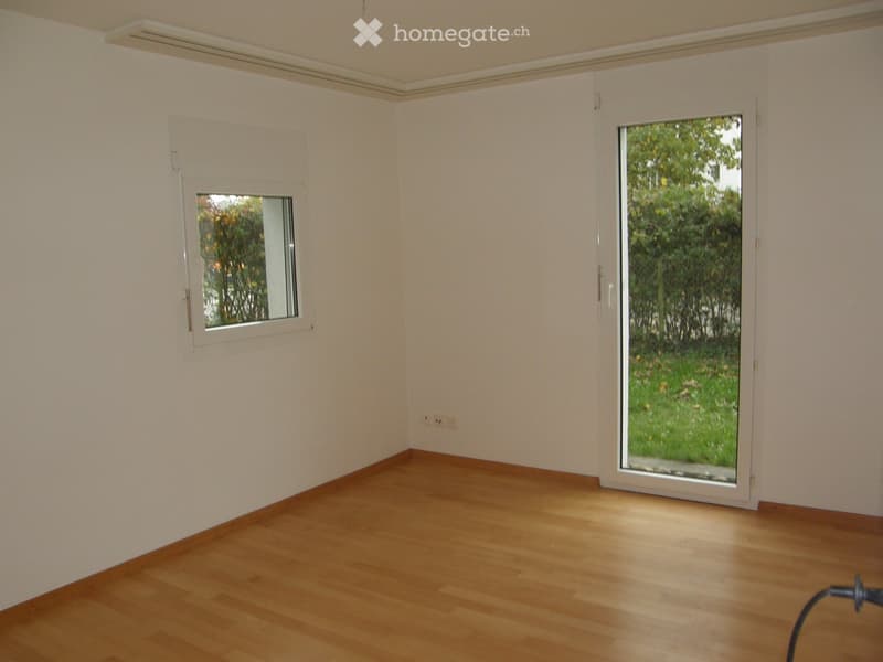 Empty room with light-colored walls, wooden flooring, and two windows, one of which is a sliding glass door opening to a green outdoor area.