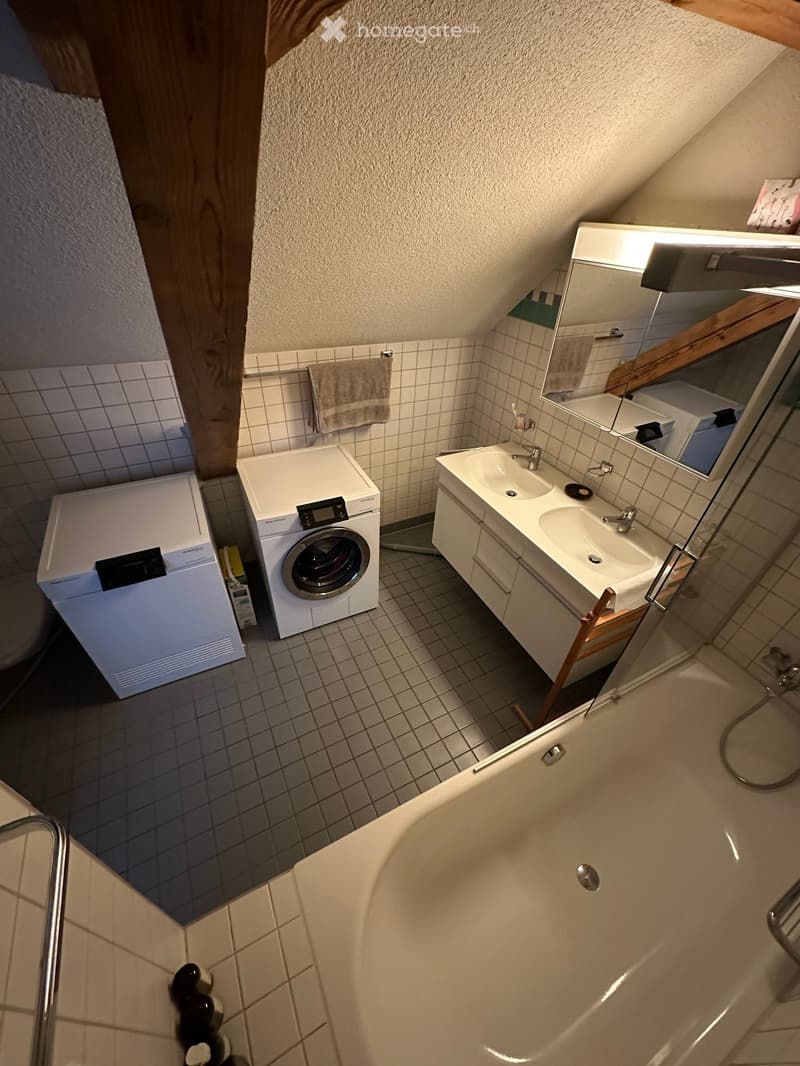Bathroom with a bathtub, sink, mirror, washing machine, and towel. The room features tiled walls and a slanted ceiling typical of an attic space.