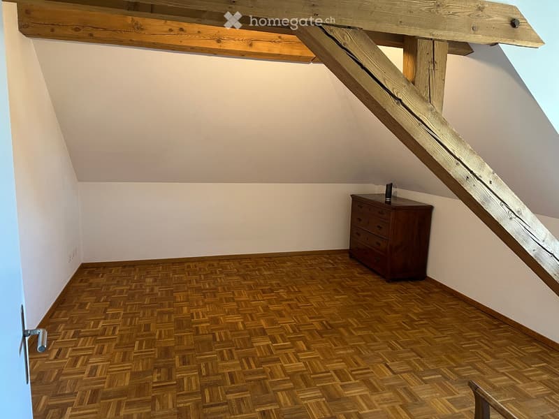 Attic room with wooden herringbone flooring, slanted wooden ceiling beams, and a small wooden chest of drawers near the staircase.