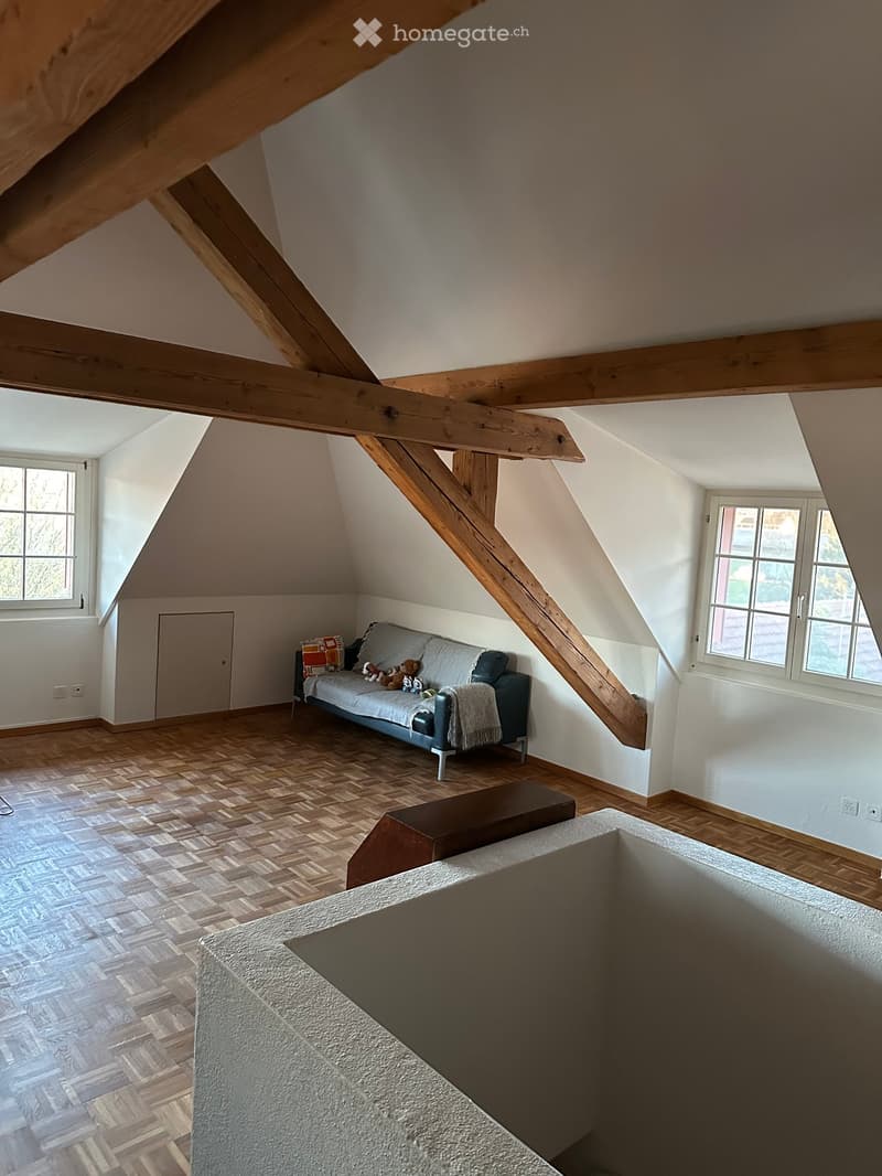 Attic room with wooden beams, slanted ceilings, and large windows. Features a grey sofa with pillows and toys, a small wooden table, and a textured white rug on the floor.