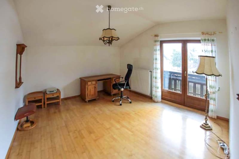 Bright room with wooden flooring, large window with balcony access, desk, chair, table lamp, fan, and mirror on the wall.