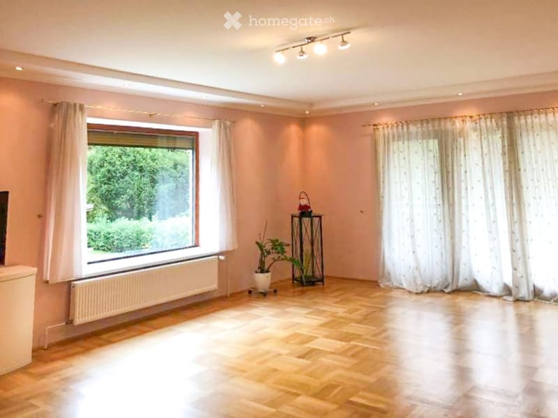 Empty room with wooden flooring, large window with white curtains, radiator, and small decorative items near the window.