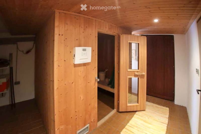 A wooden-paneled hallway with a sauna door featuring a small window, a thermostat on the wall, and a partially visible sauna interior with wooden benches and a bucket.