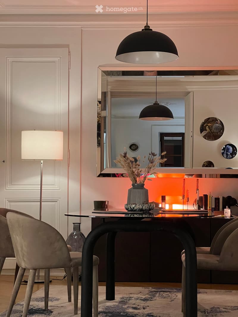 Modern dining area with a glass dining table, four chairs, a vase with dried flowers, a cup, books, candles, a mirror, and a lamp. The space features a warm, inviting ambiance with soft lighting and a stylish decor.