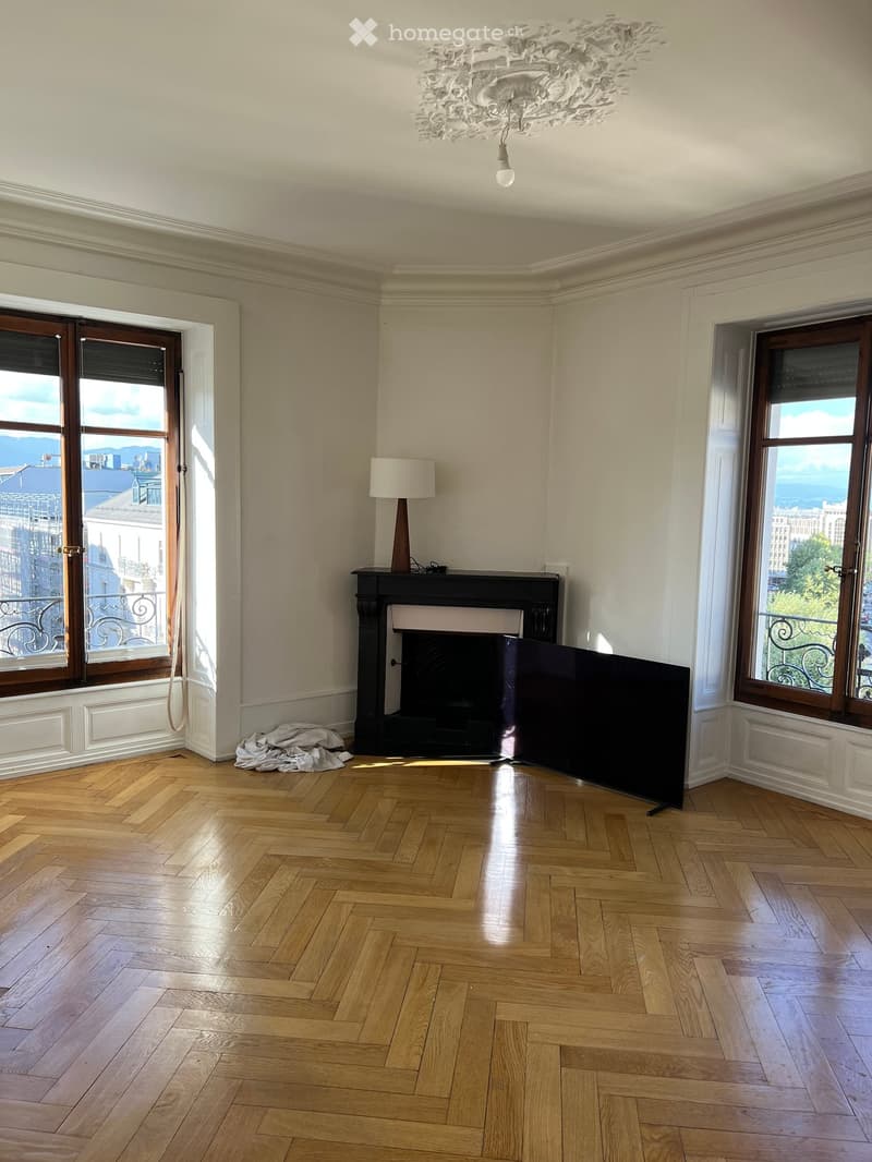 Empty room with wooden herringbone flooring, large windows offering a city view, fireplace, and decorative chandelier.